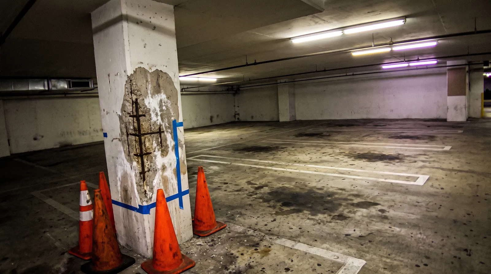 Concrete parking garage column with a spalled patch and rusty rebar, marked with blue tape and surrounded by orange traffic cones.